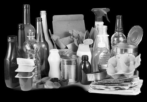 A black-and-white image showing a variety of discarded household waste items arranged on a flat surface. In the foreground, there are several glass bottles with different shapes and sizes, some with narrow necks and others with wider bodies, alongside plastic containers and disposable cups. Behind these, a cardboard box is filled with crumpled paper and other rubbish, with its flaps open, revealing more waste materials inside. To the right of the box, a pile of flattened newspapers and magazines is stacked neatly, with some loose pages on top. Several plastic spray bottles and empty food containers are also visible, along with a crumpled paper wrapping or packaging. The background appears plain and contrasts with the clutter of waste materials, suggesting a setup for rubbish collection or clearance, possibly related to a house or property cleanup in the Oxford area, as indicated by the context of House Clearance Oxford and their rubbish removal services.