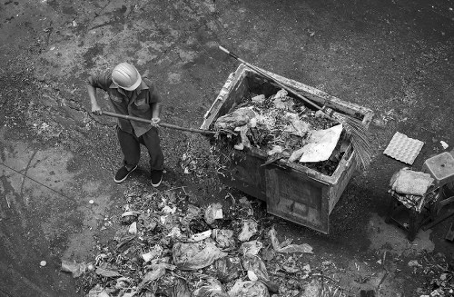 A worker wearing a hard hat and casual clothing is standing on a paved surface, using a long-handled tool to clear a pile of mixed waste and rubbish from the ground near a large, open-top metal dumpster. The rubbish includes various materials such as paper, plastic, and discarded packaging, with some waste spilling onto the pavement. The metal dumpster, positioned against a rough-textured concrete or asphalt background, is partially filled with similar debris, showing a mix of loose and compacted rubbish. The scene suggests an outdoor rubbish removal or waste clearance activity, typical of local waste management services in Oxford, with the presence of a small stack of bricks or tiles nearby. The lighting appears natural, with shadows indicating daytime, and the overall environment is utilitarian and functional, reflecting a typical scene in rubbish collection or house clearance work by House Clearance Oxford, serving areas including Oxford and postal code OX.