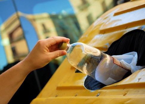 A close-up of a person's hand holding a transparent plastic bottle filled with clear liquid, preparing to dispose of it into a designated slot on a yellow waste collection point, which appears to be a recycling or rubbish bin. The surface of the bin is textured with a wooden-like finish, and the background shows a blurred view of a building and outdoor environment, suggesting the scene takes place outside. The liquid inside the bottle is visible through the clear plastic, and the person's fingers are grasping the neck of the bottle. This image emphasizes proper waste disposal practices as part of a rubbish removal service, with the setting likely in Oxford or nearby areas, as referenced in local service context. The lighting is natural, highlighting the details of the bottle and the textured surface of the bin, supporting environmentally responsible rubbish collection efforts facilitated by House Clearance Oxford.
