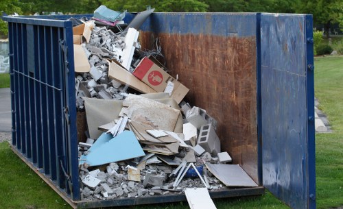 A large industrial skip container, situated outdoors on a grassy area, filled with a mixture of construction and household rubbish. The interior of the skip reveals a rusted metal surface, with its sides painted in dark blue. The contents include broken concrete slabs, chipped plasterboard, scattered cardboard boxes, pieces of wood, and assorted debris, some leaning against the sides and others piled at the base. The debris is irregularly shaped and varied in texture, with rough concrete and smooth cardboard surfaces clearly distinguishable. Environmental greenery is visible in the background, suggesting the skip is placed in an open space, possibly for a house clearance or renovation project. The presence of items like drywall and cardboard indicates non-hazardous waste. This image illustrates the typical contents handled by a professional rubbish removal service such as House Clearance Oxford, serving local areas including Oxford and postcode regions associated with the town.