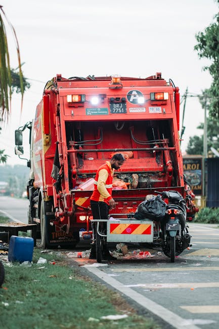 A daytime scene on a narrow urban street shows a waste collection truck with a predominantly white body and a rusty, open rear loading area, positioned parallel to the sidewalk. The truck's rear compa