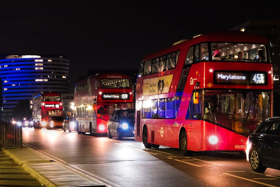 A nighttime city street scene featuring three red double-decker buses lined up in a queue, with illuminated interior lighting revealing seated passengers through large front and side windows. The fron