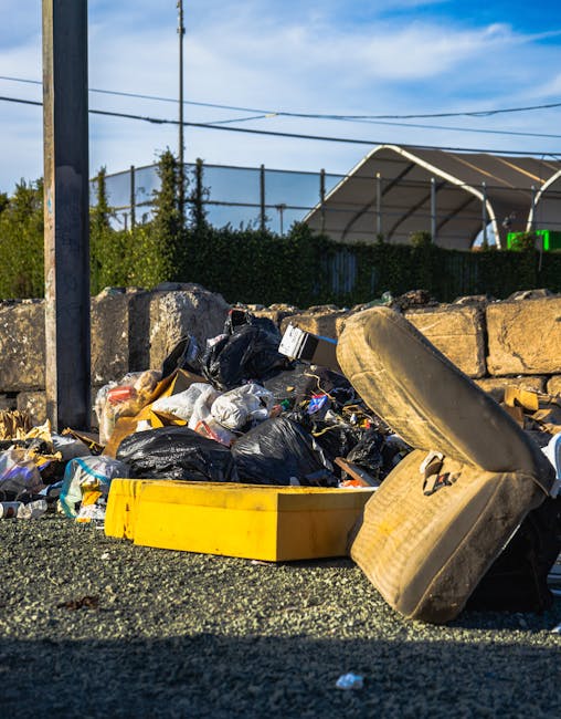 A sanitation worker in a fluorescent yellow and orange reflective uniform is seen standing at the rear of a large red waste collection truck, which is positioned on a paved urban road. The worker appe